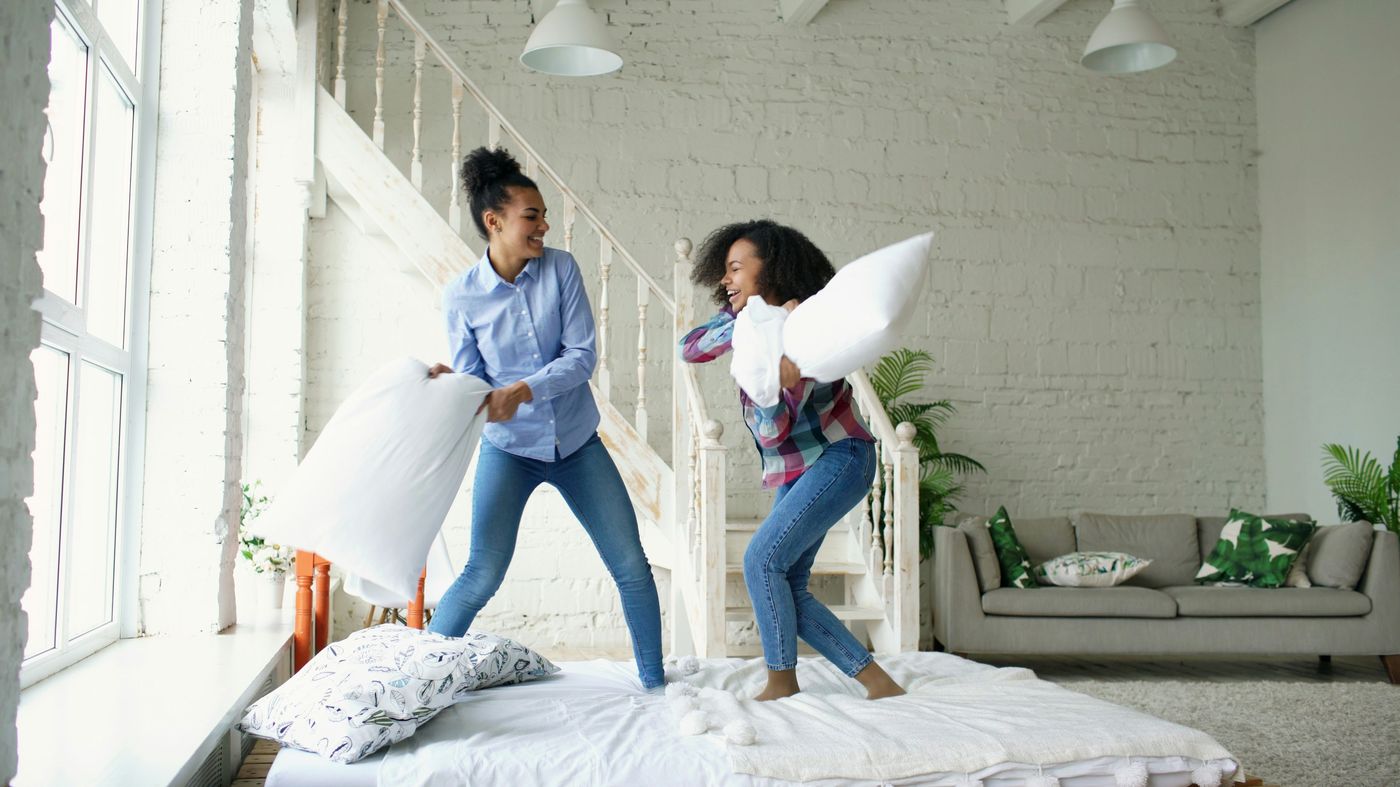 Two people celebrating with a pillow fight in the bright, freshly made bedroom of a new home